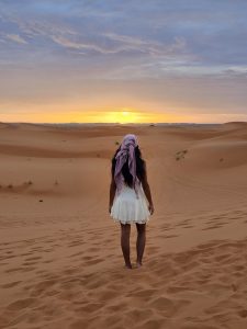 Woman in white dress walks on desert dunes at sunset.